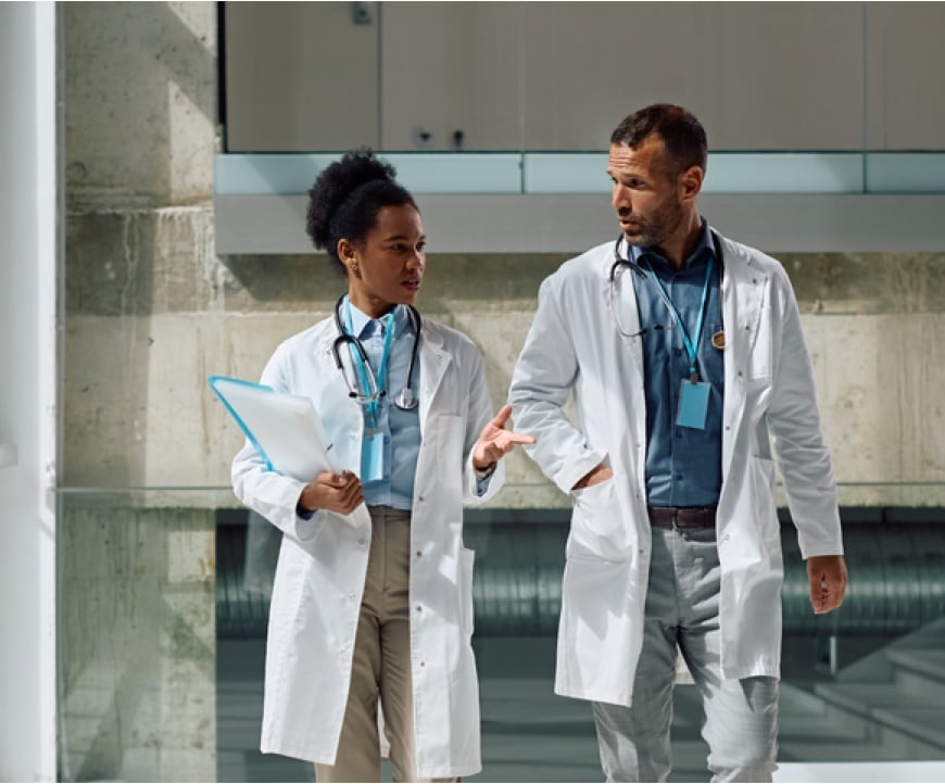 Two doctors in lab coats walk and talk, one holding a clipboard, in a modern hallway.