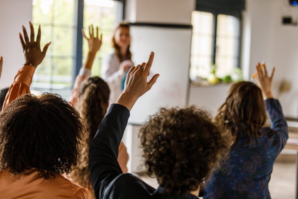 Diverse audience raising hands in a bright classroom, with a woman presenting at a whiteboard.